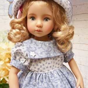 Könnte beinhalten: Eine Porzellanpuppe mit blonden Locken, die ein blau-weiß gemustertes Kleid und einen blau-weiß gemusterten Hut mit einem rosa Band trägt.