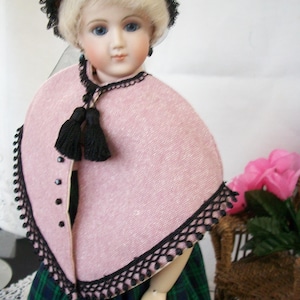 May include: A vintage porcelain doll with blonde hair and blue eyes, wearing a pink cape with black trim and tassels. The doll is dressed in a green and blue plaid skirt and a black lace bonnet. A classic collectible.