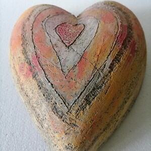 Può includere: Un oggetto decorativo a forma di cuore con un aspetto strutturato e invecchiato. Il cuore presenta forme di cuore concentriche nei toni dell'arancione, del rosa e del bianco, delineate da linee scure. L'effetto complessivo è rustico e artistico.