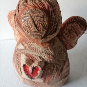 May include: A handmade ceramic angel figurine with a heart-shaped detail. The angel is primarily brown with reddish-brown wings and a red heart. The ceramic has a textured surface, and the wings are slightly spread.