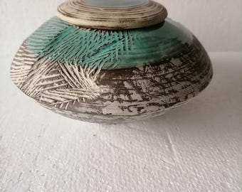 May include: A handcrafted ceramic vessel with a lid. The pottery features a textured surface with a teal and brown color scheme. The lid is a lighter shade of brown with concentric circle detailing. The vessel is a decorative piece.