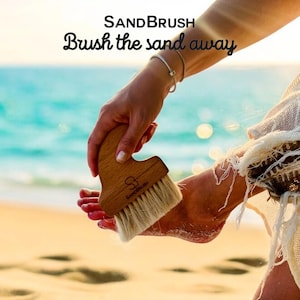 Könnte beinhalten: Eine Bürste aus Holz mit natürlichen Borsten zum Entfernen von Sand von den Füßen. Die Bürste hat einen Griff mit der Aufschrift "SandBrush". Die Bürste wird auf dem Fuß einer Person an einem Sandstrand verwendet.
