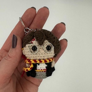 Może przedstawiać: Breloczek Harry Potter wykonany na szydełku z brązowymi włosami, okrągłymi okularami, czerwono-żółtym szalikiem i czarną szatą.
