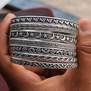 Puede incluir: Una pila de brazaletes plateados con diseños intrincados. Los brazaletes presentan patrones de pájaros, olas, lunas, estrellas y motivos florales. Los brazaletes se sostienen en una mano, mostrando la artesanía detallada.