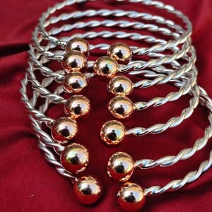 Pode incluir: Uma coleção de pulseiras de metal prateado e torcido com detalhes esféricos dourados. As pulseiras estão dispostas sobre um tecido vermelho escuro, mostrando seu design intrincado e cores contrastantes. Os detalhes esféricos adicionam um toque de elegância.