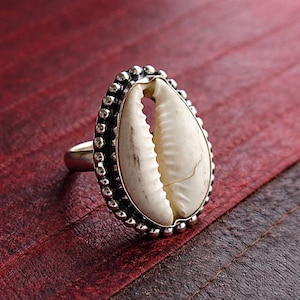Puede incluir: Un anillo de plata con una gran concha de cauri blanca. La concha está engastada en un bisel de plata con un borde de cuentas. El anillo se muestra sobre una superficie roja, mostrando el diseño y la textura de la joya.