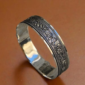 May include: A silver-colored bangle bracelet with an intricate floral and leaf pattern. The detailed design wraps around the entire band, creating a textured surface. The bracelet is displayed on a brown surface.
