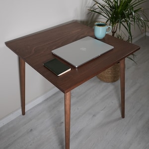Puede incluir: Una mesa auxiliar de madera marrón oscuro con cuatro patas cónicas. Una computadora portátil plateada, un pequeño cuaderno verde y una taza azul claro están sobre la mesa. Una planta en maceta está cerca. La mesa está sobre un suelo gris claro.