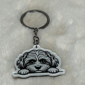 Może przedstawiać: Biały brelok akrylowy z czarno-białym rysunkiem psa wyglądającego zza krawędzi. Pies ma puszyste futro i duże oczy.