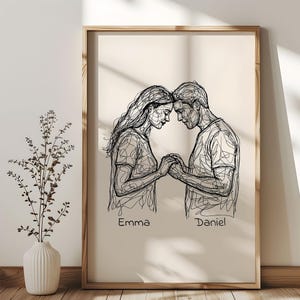 May include: A framed line art print featuring a couple holding hands, with their heads touching. The artwork is in black lines on a beige background. The names "Emma" and "Daniel" are written below the figures. The frame is made of light brown wood.