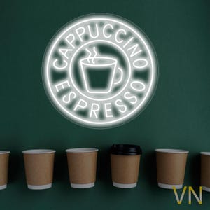 May include: A white neon sign with the words "Cappuccino Espresso" and a stylised image of a coffee cup with steam coming from it. The sign is hanging on a green wall above six brown paper coffee cups.