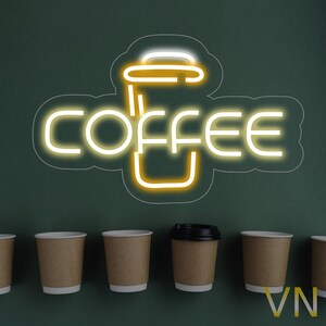 May include: A neon sign displaying the word "COFFEE" and a coffee cup outline in bright yellow against a dark green backdrop. Several brown paper coffee cups are positioned below the sign, one with a black lid.