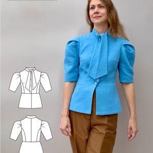 Könnte beinhalten: Eine hellblaue Bluse mit Stehkragen, kurzen Ärmeln und einem Bindedetail. Die Bluse wird mit braunen Hosen kombiniert. Eine Strichzeichnung der Bluse ist ebenfalls abgebildet. Der Markenname "EKATOL" ist sichtbar.