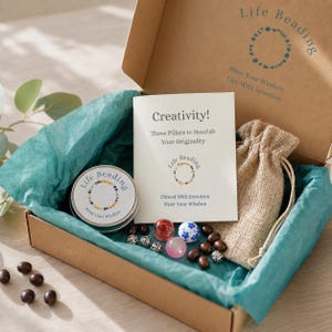 May include: A cardboard gift box containing a round metal tin with a Life Beading label, a card titled Creativity! Three Pillars to Nourish Your Originality, a small burlap drawstring pouch, and several loose beads including glass and metal pieces.