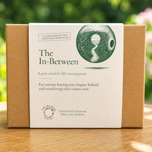 May include: A rectangular brown cardboard box with a white paper sleeve featuring the text A Guided Reflection & Bracelet-Making Kit, The In-Between, A quiet ritual for life's turning points, and a green circular illustration of a path.