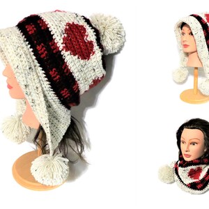 Può includere: Un cappello a uncinetto bianco, rosso e nero con un design a forma di cuore e pon pon. Il cappello ha un cappuccio lungo e appuntito ed è mostrato su un manichino.