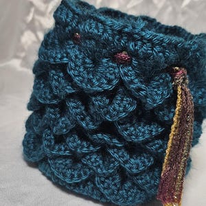 May include: Teal crochet pouch with a scale-like pattern. The handmade pouch features a decorative tassel with gold and burgundy threads. The pouch is a unique accessory, perfect for storing small items.