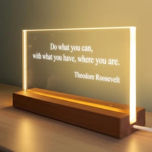 Può includere: Insegna luminosa in acrilico con la citazione "Fai quello che puoi, con quello che hai, dove sei" di Theodore Roosevelt. L'insegna è posta su una base di legno ed emette una luce calda.