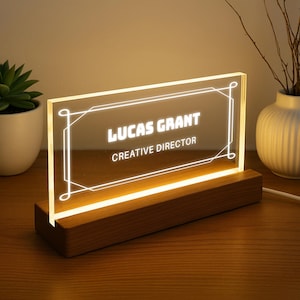 Może przedstawiać: Podświetlana tabliczka na biurko z imieniem "LUCAS GRANT" i tytułem "CREATIVE DIRECTOR" w białych literach. Prostokątna tabliczka ma ozdobną ramkę i spoczywa na drewnianej podstawie. Światło jest ciepłe.
