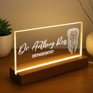 Puede incluir: Un letrero acrílico iluminado con el nombre "Dr. Anthony Ross, Orthodontist" en escritura blanca y un gráfico geométrico de un diente. Se apoya en una base de madera, emitiendo una luz cálida.