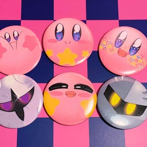 May include: Six round, colorful buttons featuring various character designs. The buttons are primarily pink, with other colors including yellow, purple, and blue. The designs include stars, faces, and other cartoon elements.