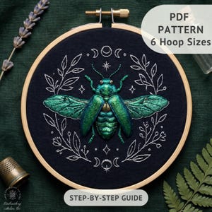 May include: An embroidery hoop containing dark fabric with a stitched iridescent green beetle centered within a white embroidered wreath of leaves and moon phases. Text on the image reads PDF PATTERN 6 Hoop Sizes and STEP-BY-STEP GUIDE.