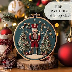 May include: Embroidered hoop art featuring a Nutcracker standing between two Christmas trees on a teal background. The hoop is made of wood and has a white snow-covered base. The text "PDF PATTERN + 6 hoop sizes" is displayed in the upper right corner of the image.