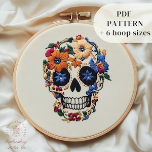 May include: Embroidered floral skull design on a white fabric in a wooden embroidery hoop. The skull is surrounded by colorful flowers in shades of orange, yellow, blue, red, and green. The text "PDF PATTERN + 6 hoop sizes" is displayed above the hoop.