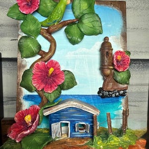 May include: A three-dimensional wall art piece featuring a green parrot perched on a brown branch with large green leaves and bright pink hibiscus flowers. Below, a blue wooden house sits near a brown stone tower and a small wire fence.