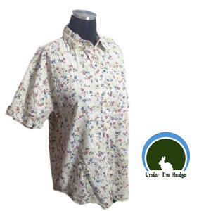 May include: A short-sleeved button-down shirt in an off-white fabric featuring a repeating floral pattern of small red and blue flowers with green leaves. The shirt has a standard collar and a front button placket. A logo with a rabbit and the text Under the Hedge is visible.