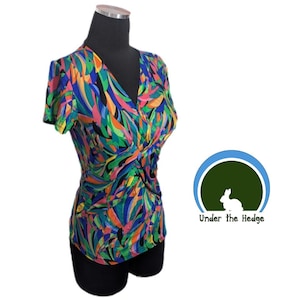 May include: A patterned short-sleeved top with a V-neck and a twisted knot detail at the waist. The fabric features an abstract design in blue, green, pink, orange, and black. The top is displayed on a black mannequin.