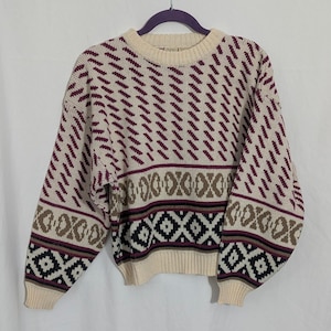 May include: Cream-colored sweater with a geometric pattern in burgundy, brown, and black. The sweater has a crew neck and long sleeves. The design includes diagonal lines and diamond shapes, creating a vintage aesthetic.