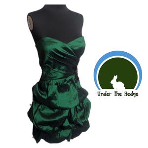 May include: A strapless, emerald green dress with a sweetheart neckline and a layered skirt. The dress is displayed on a black mannequin. A small black embellishment is at the waist. The logo "Under the Hedge" is in the background.