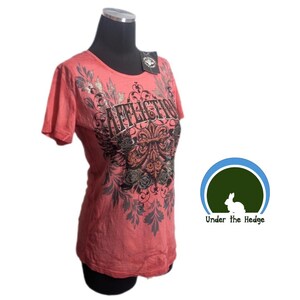 Może przedstawiać: Koralowy t-shirt z krótkim rękawem z czarnym i metalicznym nadrukiem graficznym z przodu. Wzór zawiera słowo AFFLICTION stylizowaną czcionką, otoczone skomplikowanymi motywami kwiatowymi i ornamentami. Dołączona jest czarna metka.