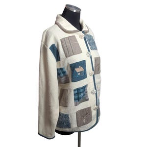 May include: A cream-colored long-sleeve jacket featuring a patchwork design with various blue, gray, and brown square panels. The jacket includes embroidered details of a small house and branches, a fold-over collar, and round buttons down the front.