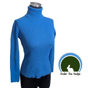 May include: A bright blue long-sleeve ribbed turtleneck sweater displayed on a mannequin. Next to the sweater is a circular logo featuring a white rabbit silhouette on a dark green background with the text Under the Hedge in black lettering.