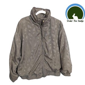 May include: A taupe-colored zip-up jacket featuring a repeating pattern of outlined leaves. The jacket has a high collar, long sleeves with elastic cuffs, and an elasticized hem. A circular logo with a white rabbit and the text Under the Hedge is visible.