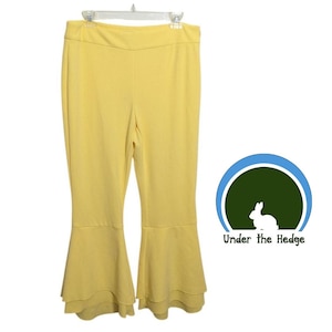 May include: Yellow flared trousers hanging on a white hanger. The trousers have a high waist and a double-layered flare at the bottom. The trousers are made of a soft, flowing fabric. The logo "Under the Hedge" is in the bottom right corner.