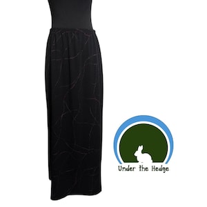 May include: A black maxi skirt featuring a subtle, thin, swirling red line pattern throughout the fabric. The skirt has an elasticized waistband and a straight, floor-length silhouette. A logo with a rabbit and the text Under the Hedge is visible.