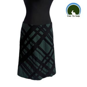 May include: A knee-length skirt in a dark forest green hue featuring a prominent black plaid pattern with diagonal lines. The garment is displayed on a black mannequin torso. A circular logo in the top right corner depicts a white rabbit with the text Under the Hedge.