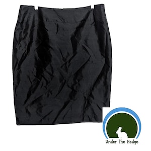 May include: A black pencil skirt made of a smooth, possibly synthetic fabric. The skirt has a straight cut and appears to be knee-length. The skirt is hanging on a white hanger. The logo "Under the Hedge" is in the bottom right corner.