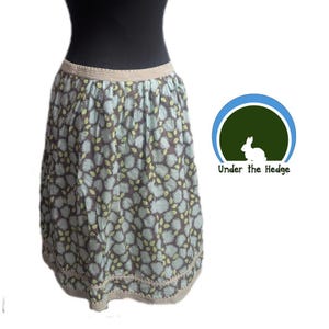 May include: A knee-length skirt with a floral pattern in shades of gray, blue, and yellow. The skirt has a crocheted waistband and hem. The brand logo "Under the Hedge" is visible on the right side.