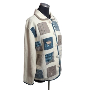 May include: Cream-colored jacket with a patchwork design featuring squares of blue, gray, and patterned fabrics. The jacket has a rounded collar, button closures, and embroidered house and nature motifs. The jacket is displayed on a mannequin.