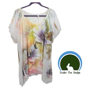 Puede incluir: Una blusa de manga corta con estampado floral en tonos blancos, amarillos, rosas y morados. La blusa está colgada de una percha negra. El estampado floral es grande y cubre toda la parte delantera de la blusa.