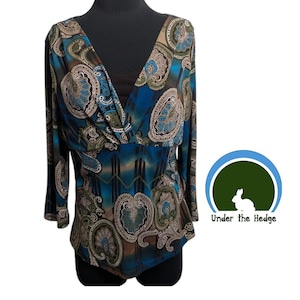 May include: A long-sleeved top featuring a deep V-neckline and a wrap-style bodice. The fabric displays a vibrant paisley pattern in shades of blue, brown, and cream, accented by geometric lines. A logo with a rabbit and the text Under the Hedge.
