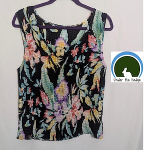May include: Sleeveless black top with a vibrant floral pattern. The top features a textured fabric and a scoop neckline. The floral design includes pink, yellow, purple, and blue flowers with green leaves. The top is hanging on a purple hanger.