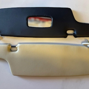 May include: Two car sun visors, one black and one beige, with a small rectangular window in the center of each. Both visors have a small round hole on the side and a white plastic clip on the back.