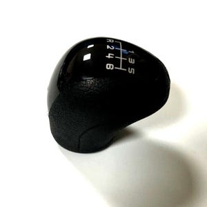 Puede incluir: Un pomo de palanca de cambios negro con una parte superior brillante y una base texturizada. El patrón de cambio está impreso en blanco, mostrando las posiciones de marcha atrás, 1, 2, 3, 4, 5 y 6. El pomo está diseñado para una transmisión manual.