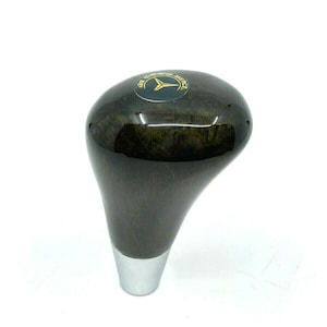 May include: A dark brown wooden gear shift knob with a silver base. The knob has a black and gold Mercedes-Benz logo on the top.