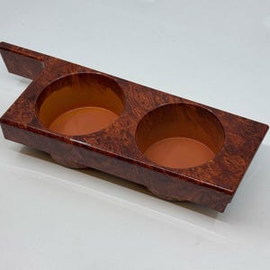 Puede incluir: Un portavasos de madera de imitación de madera marrón con dos aberturas circulares. El portavasos tiene una pequeña asa en el lateral.